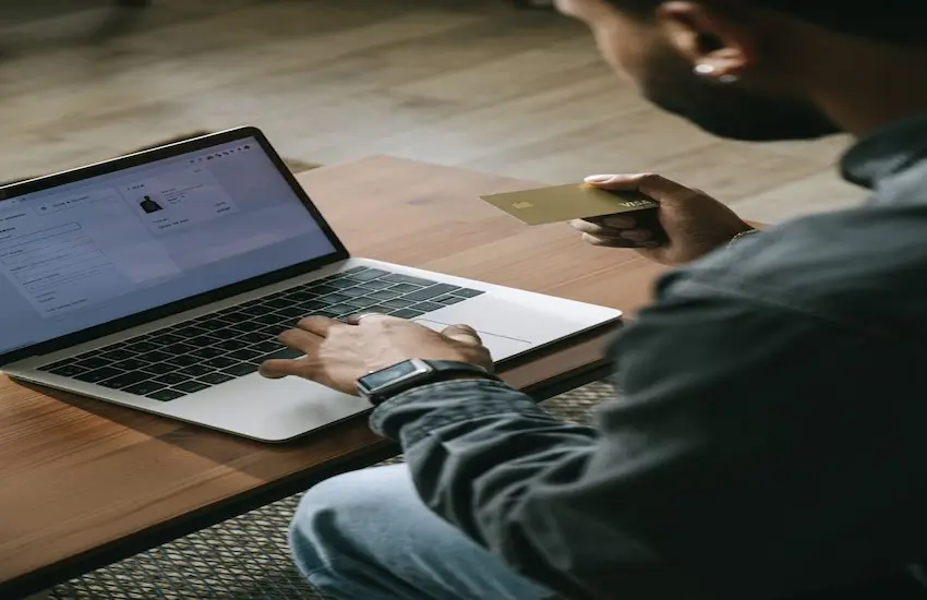 A man sitting at a desk, typing on his laptop while holding a gold credit card, analyzing personal vs business expenses on screen.