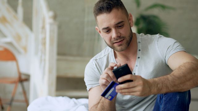 A young man sitting on a bed using a smartphone to enter credit card details for an online purchase.