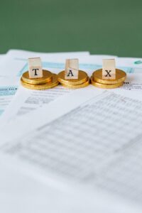 Three wooden letter blocks spelling the word "TAX" resting on small stacks of gold coins over official tax forms, illustrating how bonuses are taxed.
