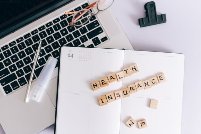 "Top-down view of an open planner on a desk with wooden letter tiles spelling 'HEALTH INSURANCE'. Next to the notebook are a laptop, eyeglasses, a pen, and a binder clip, symbolizing healthcare financial planning, deductibles, and out-of-pocket maximum calculations."