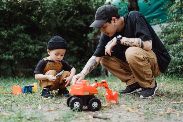 "A father and his young son crouch together on the grass, playing with a large orange toy excavator. The father is guiding the child, illustrating a strong parental bond and financial support in a nurturing environment."