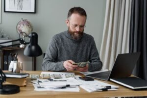 A focused man with a beard and glasses sits at a wooden desk counting stacks of cash, surrounded by financial documents, a laptop, and a smartphone, calculating his retirement savings goals.