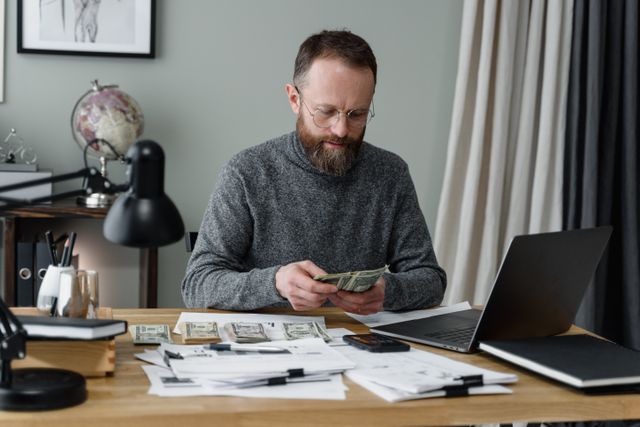 A focused man with a beard and glasses sits at a wooden desk counting stacks of cash, surrounded by financial documents, a laptop, and a smartphone, calculating his retirement savings goals.