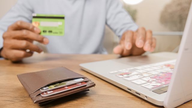 A person holding a green credit card while typing on a laptop, with a brown leather wallet filled with credit cards sitting on the wooden desk in the foreground