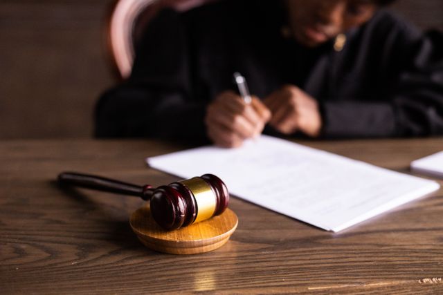 A wooden judge gavel in the foreground with a legal professional in the blurry background writing on a document.