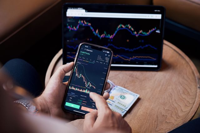 "A trader analyzing financial market charts on a smartphone and tablet next to US dollar bills, evaluating if it is a good time to invest."
