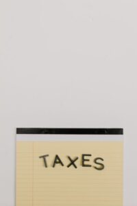 "A yellow lined legal notepad with the word 'TAXES' spelled out in dark, block letters on a plain white background."