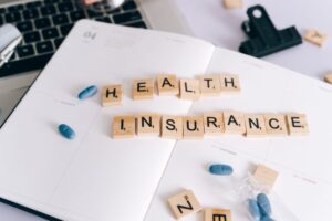 "Flat lay of a notebook with wooden letter tiles spelling 'Health Insurance' alongside blue pills and a laptop, conceptualizing self-employed health insurance costs and tax deductions."