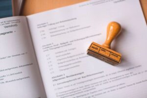"A wooden rubber stamp with the German word 'geprüft' (verified) resting on an open legal document outlining official investment regulations."
