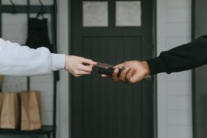 Close-up of a customer using a credit card to make a contactless payment on a handheld card reader held by a delivery person at a doorway.