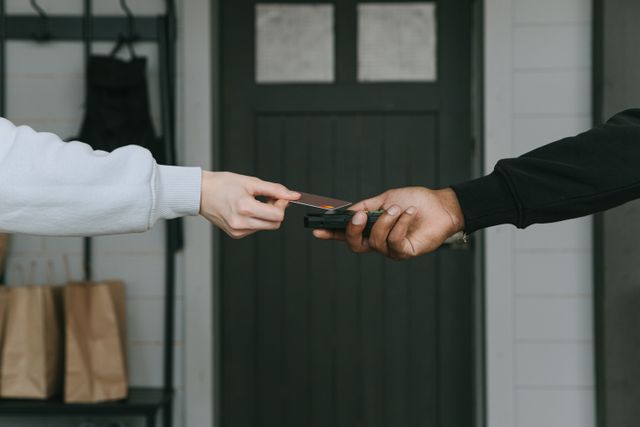 Close-up of a customer using a credit card to make a contactless payment on a handheld card reader held by a delivery person at a doorway.