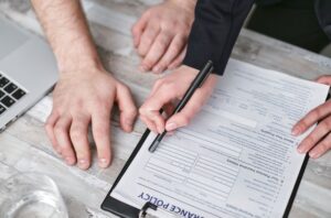 "Top-down view of a person using a black pen to point at sections of a life insurance policy form on a clipboard, while another person sits nearby, illustrating the process of reviewing complex policy terms and clauses.