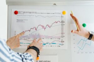 Two investors pointing at a detailed stock market trend chart pinned to a whiteboard, analyzing technical financial indicators and currency data for building a diverse investment portfolio.