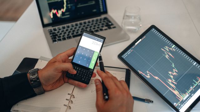 A trader analyzing stock market financial charts on a laptop and tablet while using a smartphone calculator to compute investment data over a notebook.