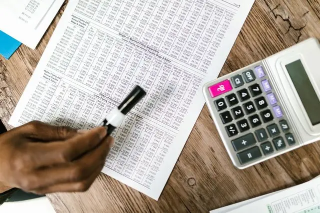 An overhead shot of a person’s hand using a black marker to review a complex financial document with numerical data tables on a wooden desk next to a digital calculator, illustrating the manual process of calculating fees and returns compared to automated robo-advisors.