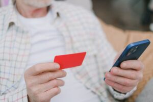 An older man holding a red credit card in one hand and looking at his smartphone, trying to resolve a denied credit card application.