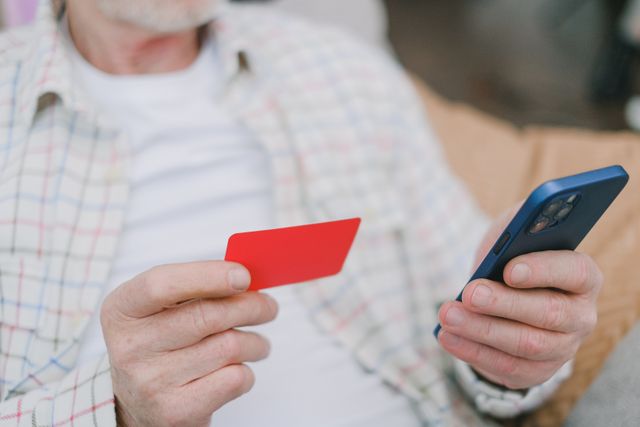 An older man holding a red credit card in one hand and looking at his smartphone, trying to resolve a denied credit card application.