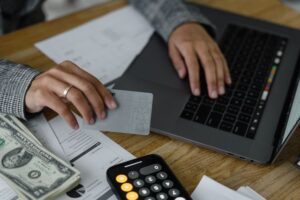 A close-up view of a person using a laptop while holding a silver credit card, with a stack of US dollar bills, financial documents, and a calculator phone application on a wooden desk.