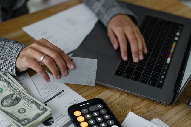 A close-up view of a person using a laptop while holding a silver credit card, with a stack of US dollar bills, financial documents, and a calculator phone application on a wooden desk.