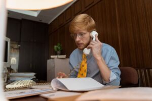 "A stressed male debt collector with glasses and a yellow tie sitting at a vintage office desk, holding a landline phone receiver and reviewing financial paperwork."
