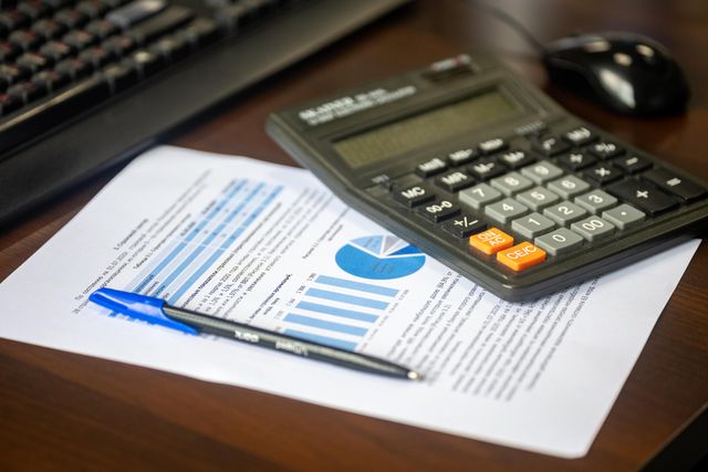 A close-up shot of a black calculator, a blue pen, and a financial document with charts on a wooden office desk.