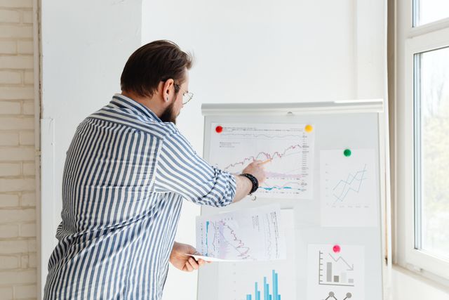A financial professional in a striped shirt points to a rising stock market trend graph on a whiteboard during a presentation about 2026 FHA loan requirements, illustrating concepts like credit score benchmarks, down payment options, and Mortgage Insurance Premium (MIP) updates.