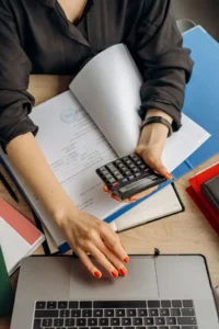 An accountant or freelancer reviewing financial documents, holding a calculator, and typing on a laptop to manage health insurance costs and budget plan without a job.
