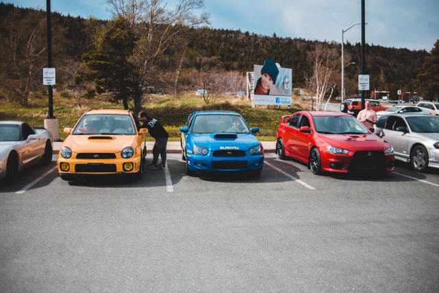 "A line of performance sports cars, including a yellow, blue, and red Subaru and Mitsubishi, parked in a lot, representing consumer spending choices on vehicles."