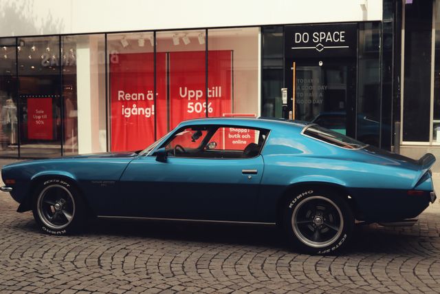 A classic metallic blue second-generation Chevrolet Camaro Z28 sports car parked on a cobblestone street in front of a modern storefront window displaying red sale signs.