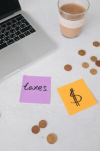 A laptop, a cup of coffee, loose coins, and sticky notes reading "taxes" and a dollar sign on a white desk.