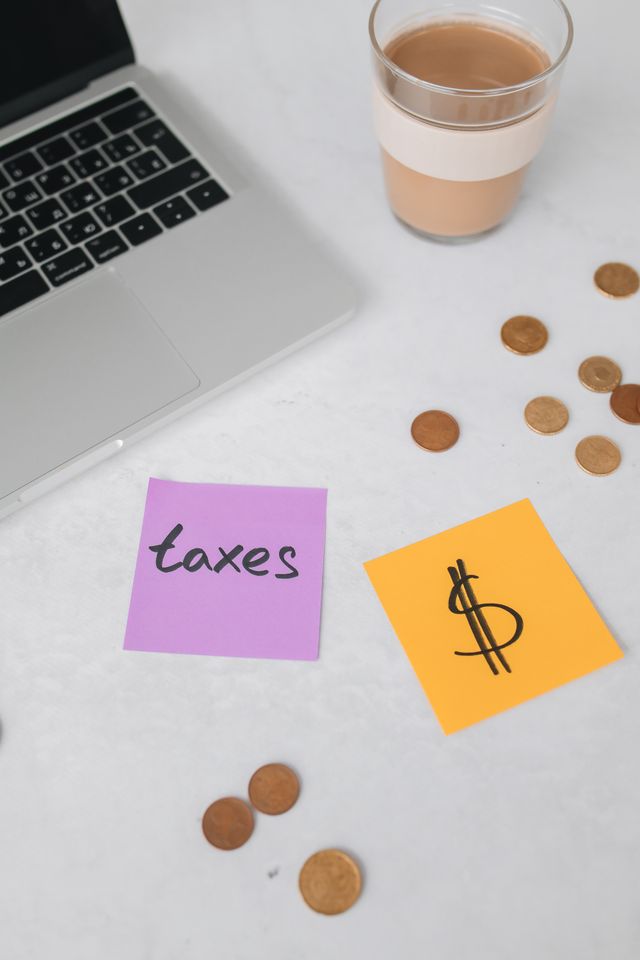 A laptop, a cup of coffee, loose coins, and sticky notes reading "taxes" and a dollar sign on a white desk.