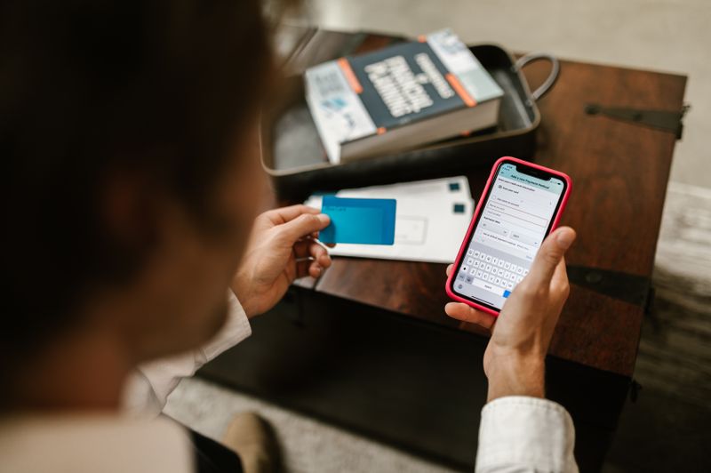 A person holds a blue credit card in one hand while using a smartphone with a red case in the other to enter financial information. In the background, a book titled "Financial Planning" sits on a wooden table alongside white envelopes.