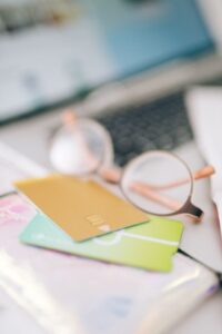 A close-up shot of a gold credit card with a visible EMV chip and contactless payment symbol, resting on top of a green card. Both cards are placed on a wallet next to a pair of glasses, with a blurred laptop and smartphone in the background, symbolizing online shopping and digital banking.