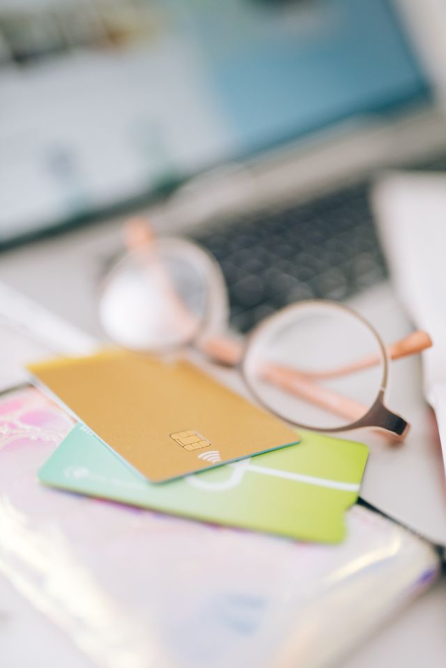 A close-up shot of a gold credit card with a visible EMV chip and contactless payment symbol, resting on top of a green card. Both cards are placed on a wallet next to a pair of glasses, with a blurred laptop and smartphone in the background, symbolizing online shopping and digital banking.