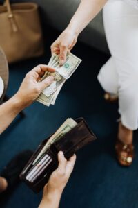 A top-down view of a person handing cash to another person, pulling US twenty-dollar bills from a brown leather wallet to represent financial transactions, budgeting, and steps to get out of debt.