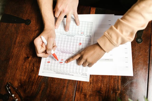 Two people reviewing a printed financial budget worksheet with rows of numbers on a wooden table, with one person pointing at a figure and the other marking data with a red pen.