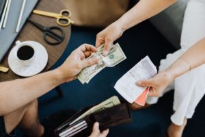 An overhead shot of a couple splitting finances, with one person handing over US dollar bills while the other holds receipts, and an open wallet visible below.
