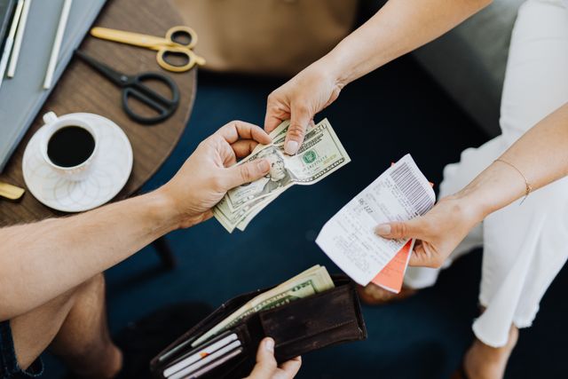 An overhead shot of a couple splitting finances, with one person handing over US dollar bills while the other holds receipts, and an open wallet visible below.