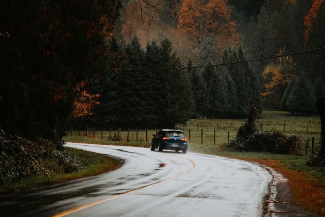 A dark hatchback car drives along a wet, winding asphalt road during autumn, showcasing safe and continuous navigation through challenging conditions.