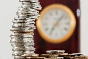 A tall stack of silver coins next to a blurred clock, illustrating the long-term impact of investment fees over time.