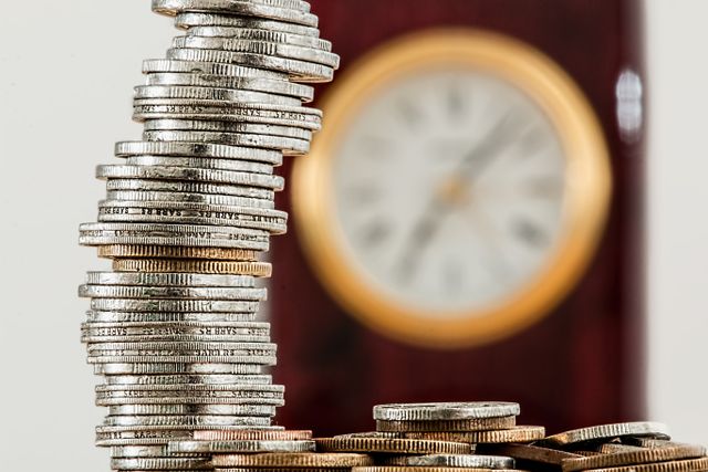 A tall stack of silver coins next to a blurred clock, illustrating the long-term impact of investment fees over time.