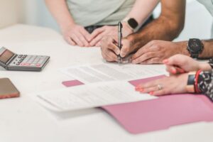 A close-up of people signing financial documents and contracts on a white desk with a calculator, smartphone, and pink folders, highlighting the process of managing payouts and policy values.
