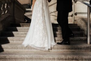 An elegant couple standing on stone stairs, showcasing a detailed lace wedding dress and a black tuxedo, illustrating the step-by-step process of obtaining a Marriage Green Card in 2026.
