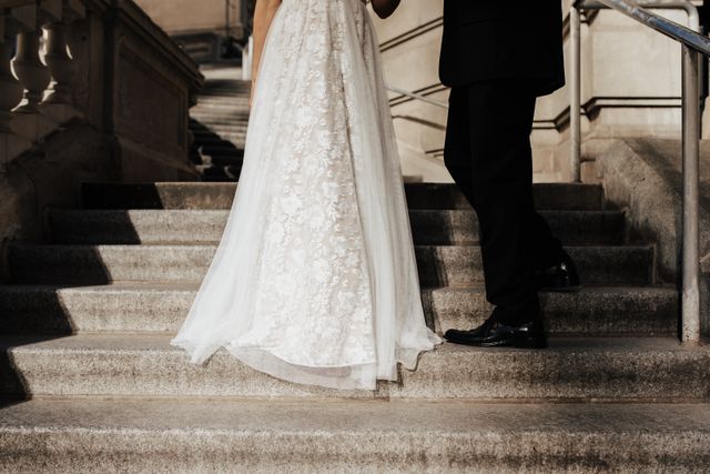 An elegant couple standing on stone stairs, showcasing a detailed lace wedding dress and a black tuxedo, illustrating the step-by-step process of obtaining a Marriage Green Card in 2026.