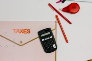 Top-down view of a light pink accordion folder labeled 'TAXES' with a small black calculator resting on it. Next to the folder are two red pens and a red correction tape dispenser on a clean white desk.