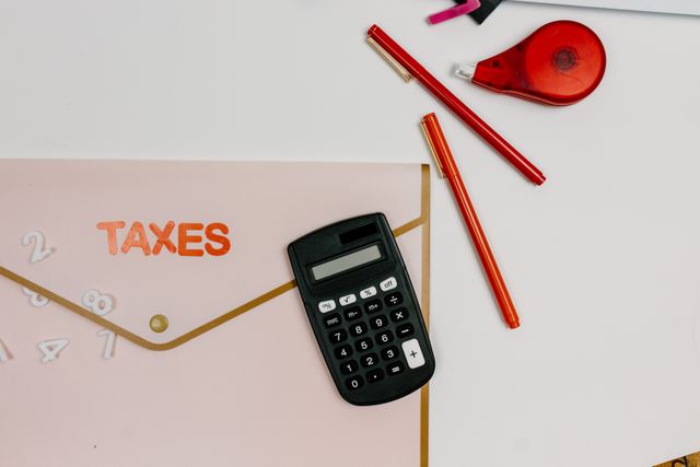 Top-down view of a light pink accordion folder labeled 'TAXES' with a small black calculator resting on it. Next to the folder are two red pens and a red correction tape dispenser on a clean white desk.
