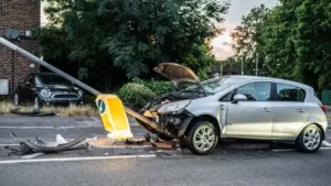 A silver hatchback car with severe front-end damage and an open hood after crashing into a yellow street signpost, demonstrating a totaled car scenario.