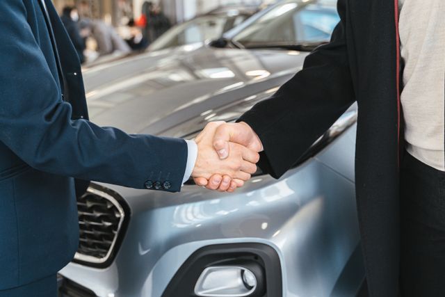 A close-up of a car salesperson in a blue suit shaking hands with a customer in front of a silver car inside a dealership showroom.