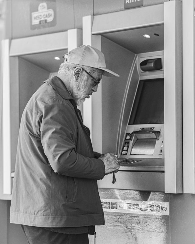 A black and white photo of a senior man wearing a baseball cap and glasses as he uses an automated teller machine (ATM).
