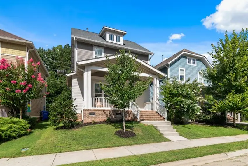 A craftsman-style, two-and-a-half-story house with a grey and white exterior, a covered front porch with white railings, and a small green lawn with a tree in front.